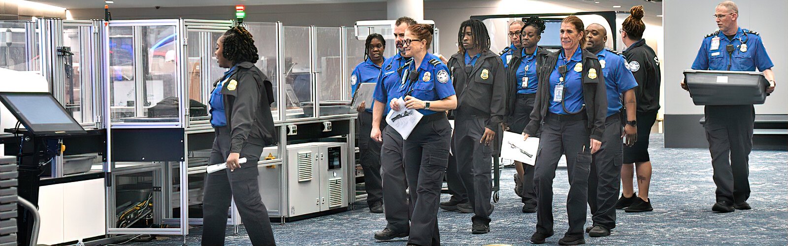  Transportation Security Officers (TSO) gather by the Airside E security checkpoint’s new automated screening lane,  where unloaded bins automatically return to the start of the lane.